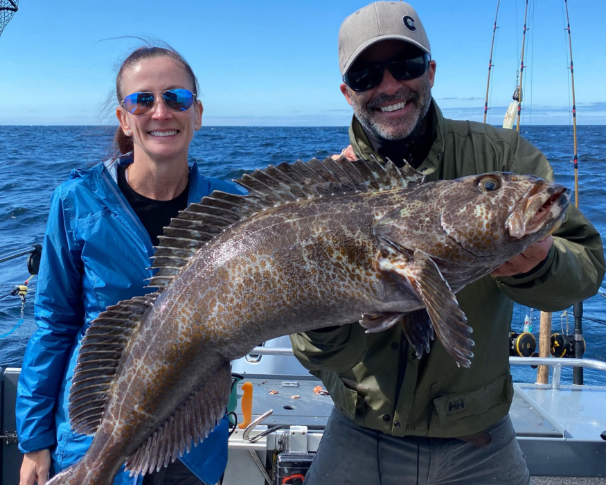 Two people standing on a boat holding a massive lingcod
