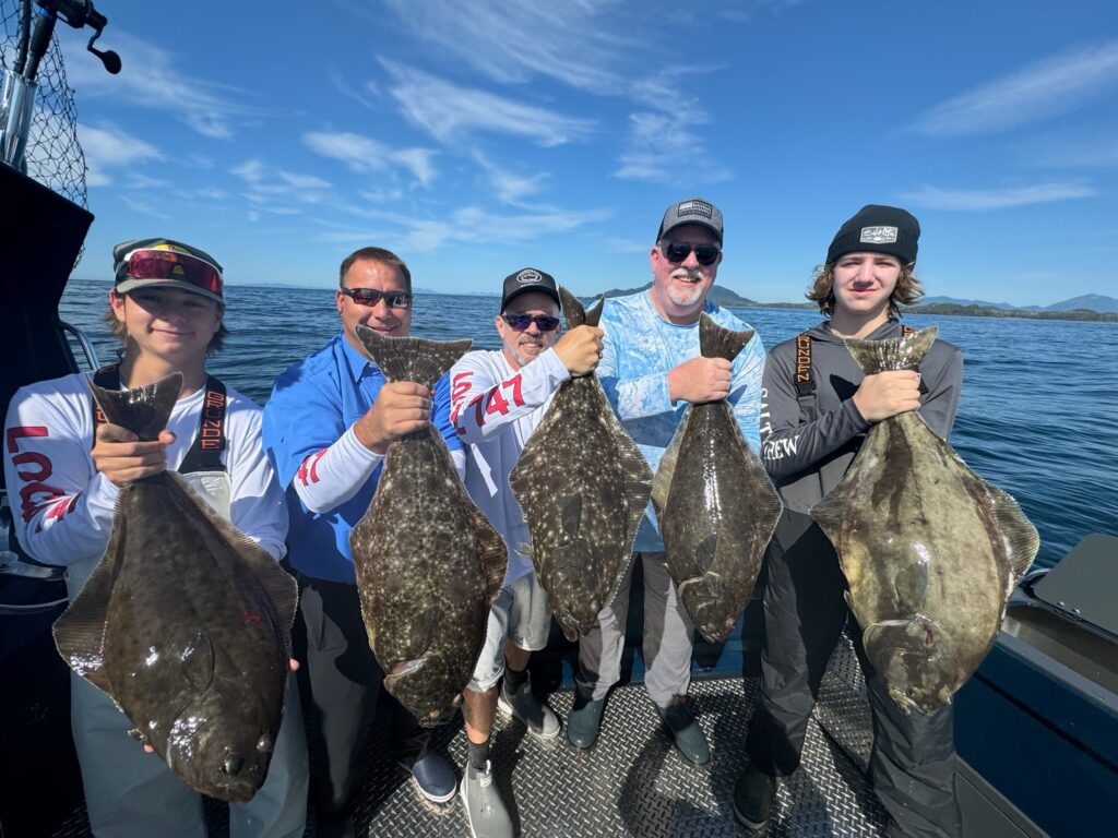 Five guys standing on a boat holding massive halibut caught during a ketchikan halibut fishing charter on a sunny day.