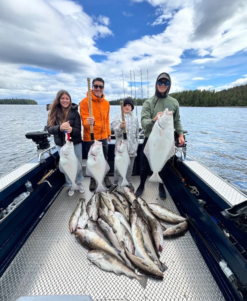 A family of 4 standing on a boat holding huge halibut, rockfish and cod on a sunny day.