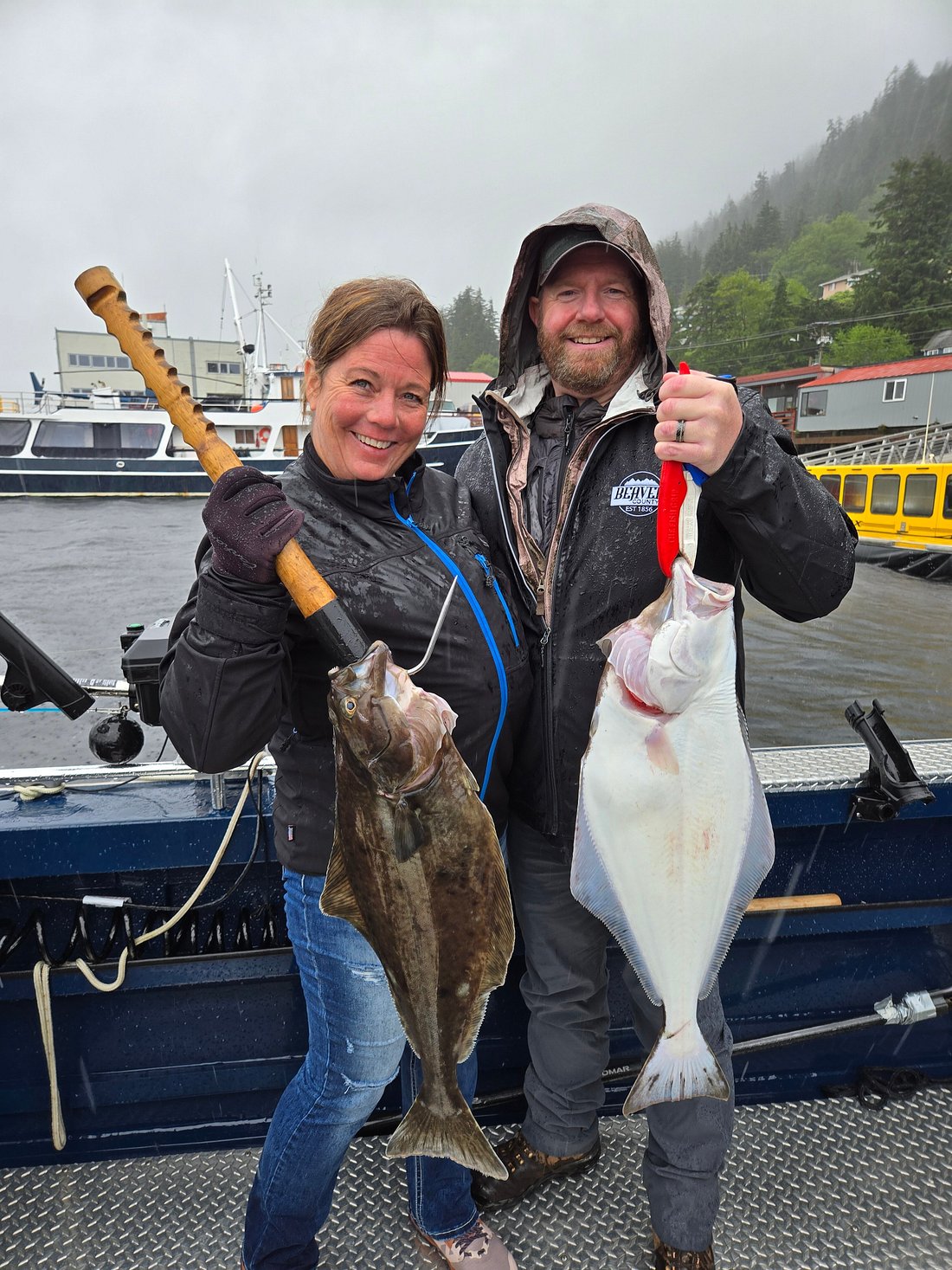 A man and a woman standing on Ketchikan finest fishing charter boat holding massive halibut with fish hooks on a cloudy, foggy day.