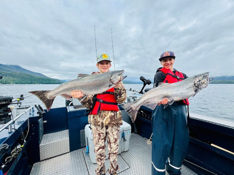 Two kids standing on a fishing charter boat on a cloudy day holding massive pacific salmon in Ketchikan during a fishing trip.